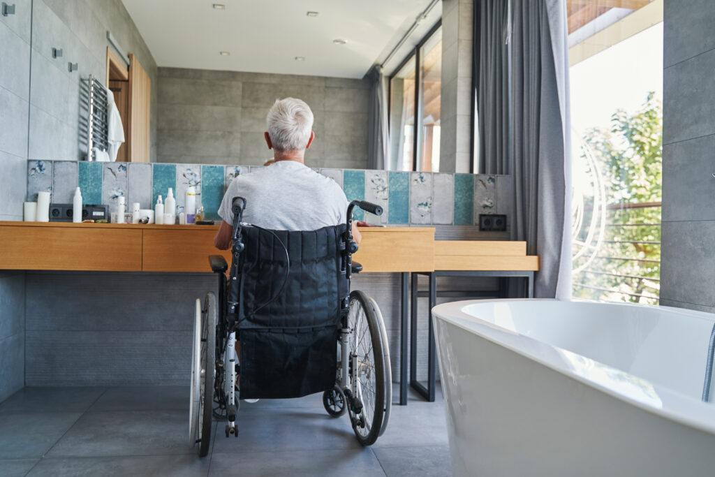 Back view of old man in wheelchair before bathroom mirror